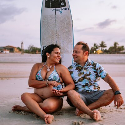 Casal sentado na areia da praia com uma prancha de surf atrás, eles se olham e sorriem eles estão segurando um par de sapatinhos de bebê e ela está grávida